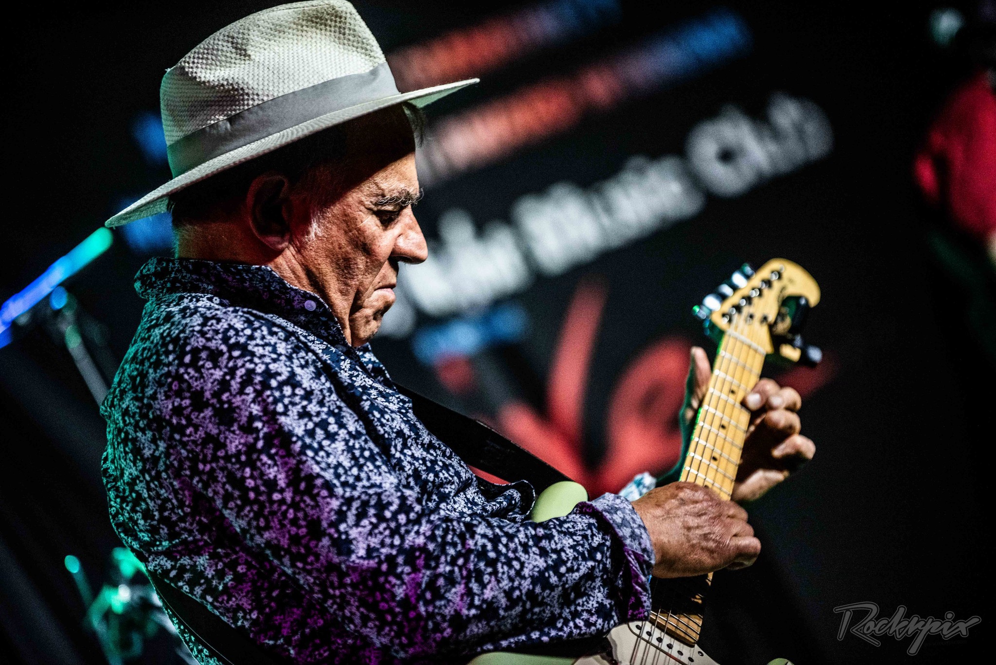 Side-profile of Robin Bibi in a white hat and patterned shirt, focused on his guitar
